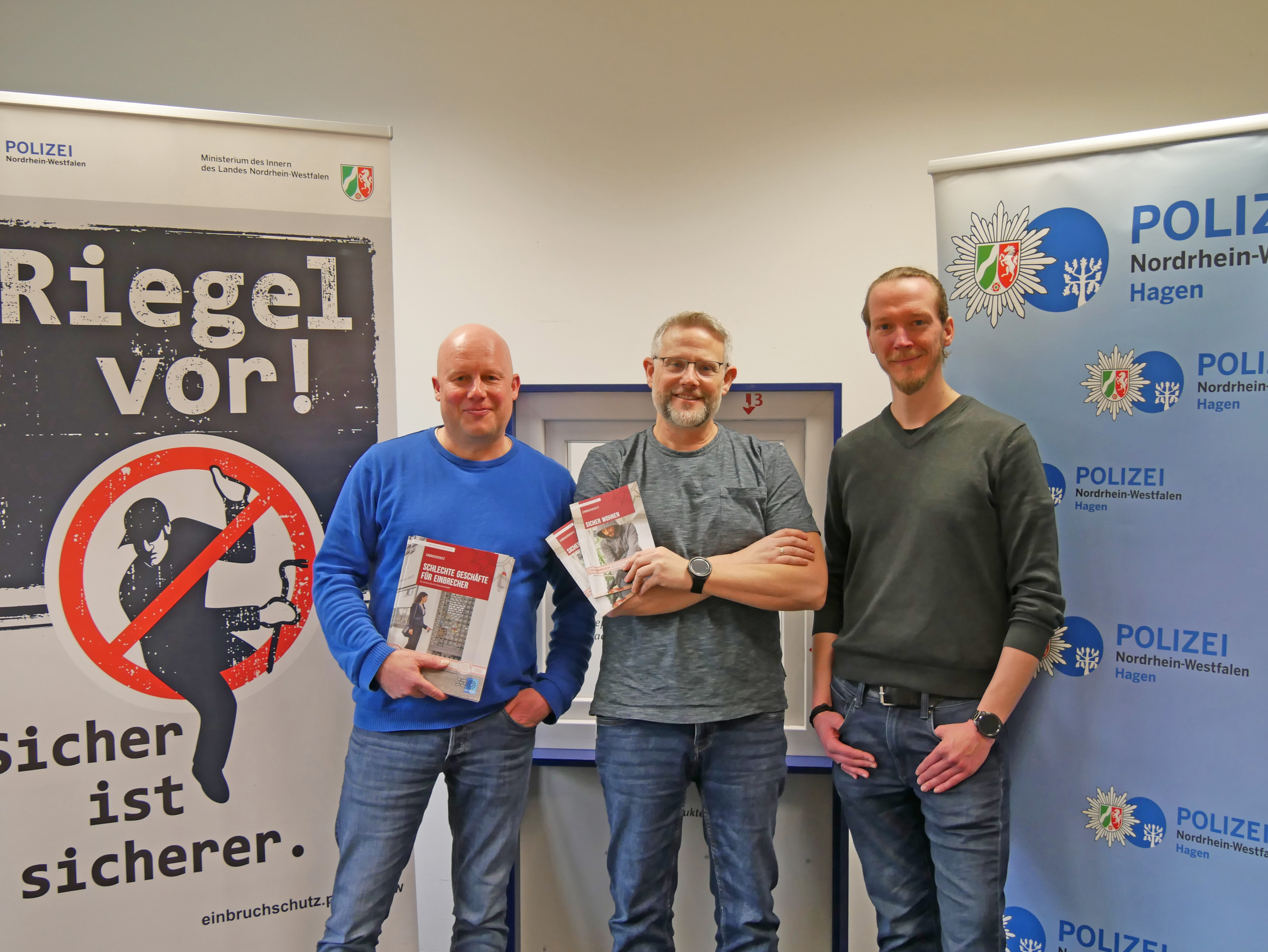 Drei lächelnde Männer stehen nebeneinander und halten Broschüren für Einbruchschutz in den Händen. Hinter ihnen befinden sich große Banner von Präventionskampagnen.