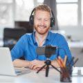 Ein junger Mann sitzt am Schreibtisch, er trägt Kopfhörer und nimmt mit seinem Smartphone ein Video auf